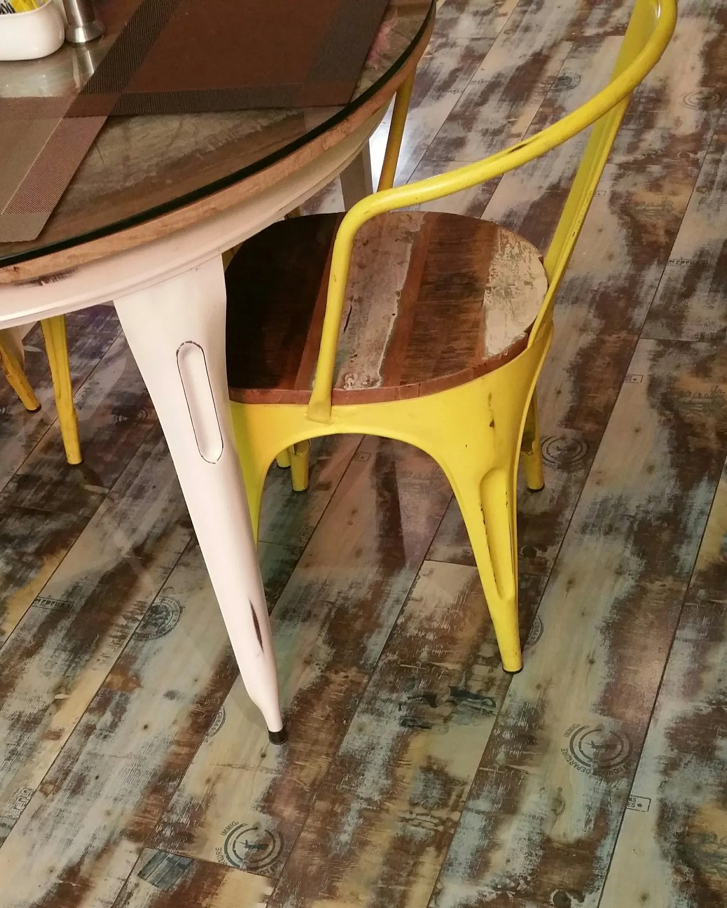 Dining area with distressed wood flooring and yellow metal chairs around a round glass-top table.