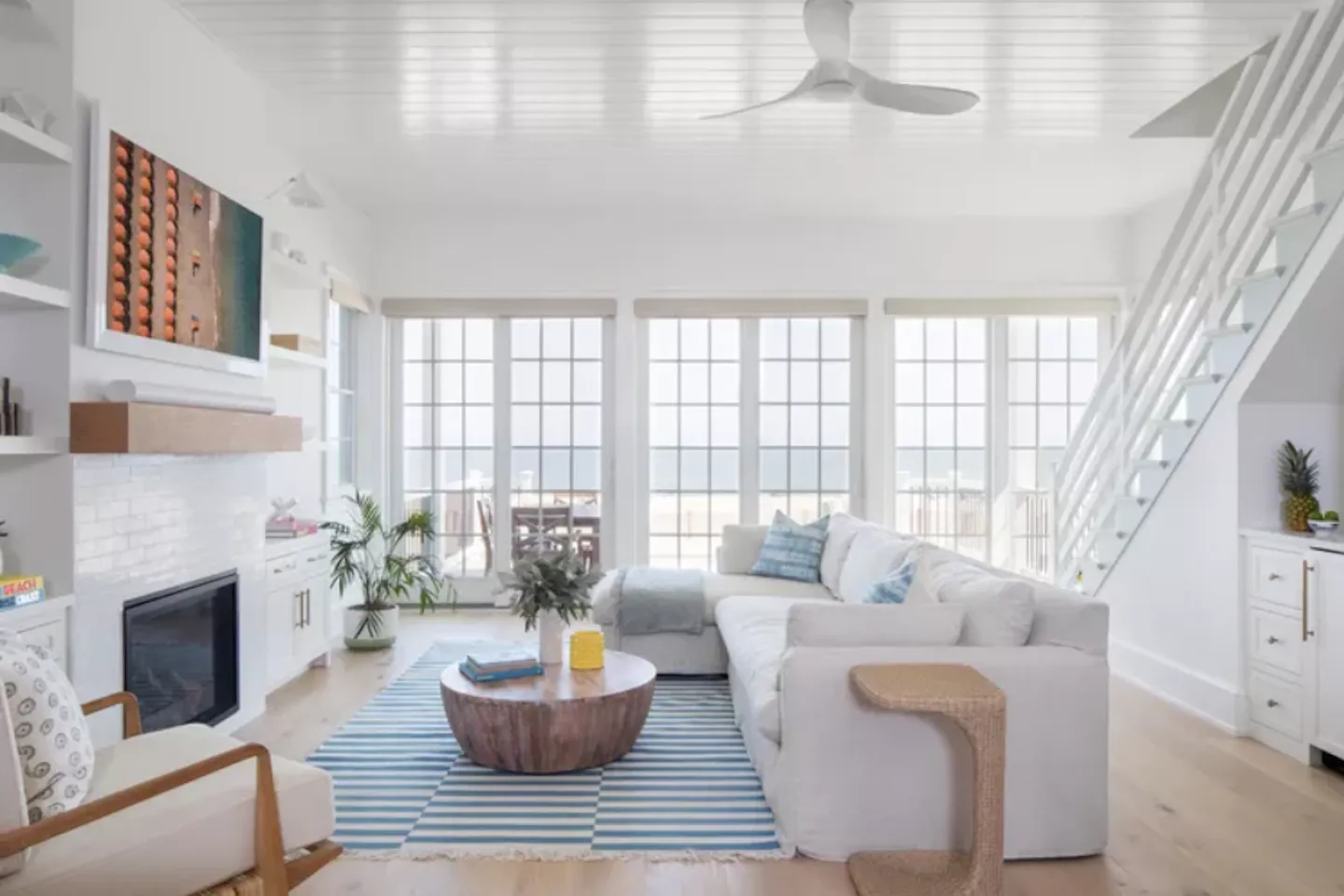 Coastal-inspired living room with light hardwood flooring, white furniture, blue throw pillows, and green plants, featuring a cozy and airy beach house decor style.